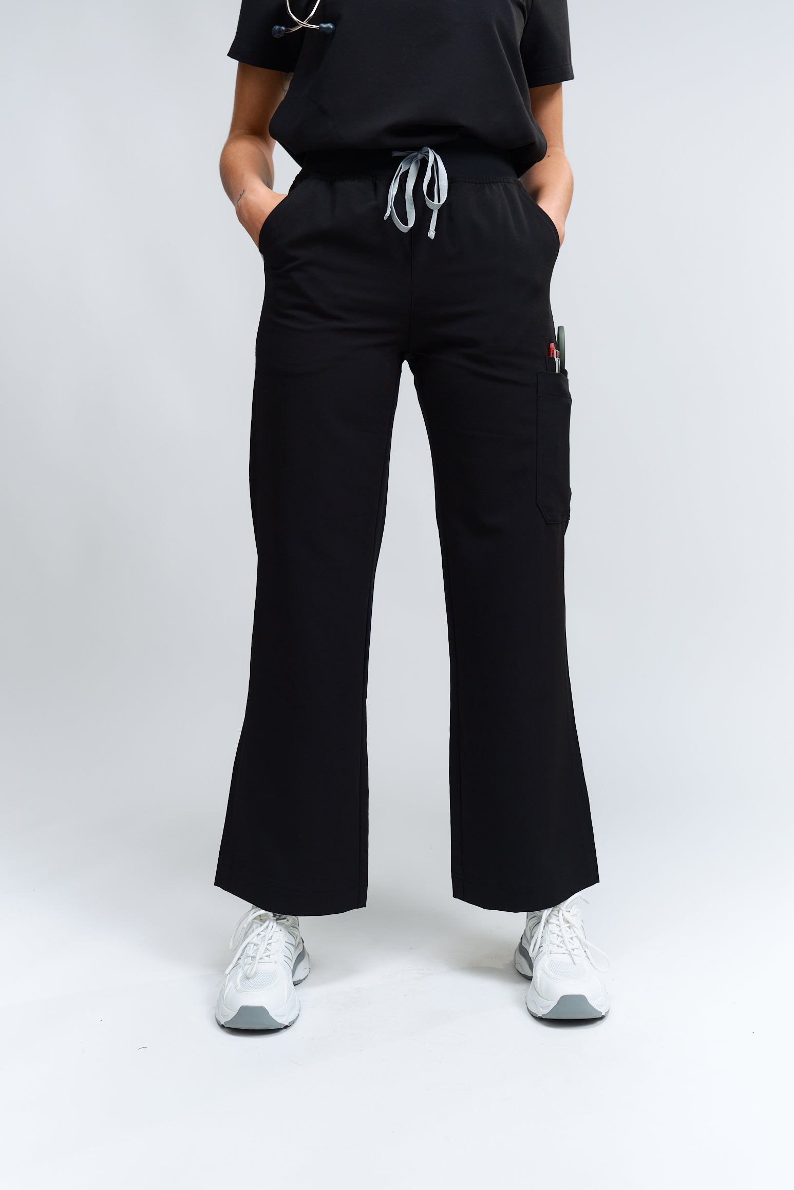 Une personne debout portant un pantalon de gommage noir à taille haute et à jambes larges, avec une ceinture élastique et un tissu antimicrobien.