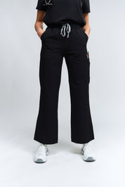 Une personne debout portant un pantalon de gommage noir à taille haute et à jambes larges, avec une ceinture élastique et un tissu antimicrobien.