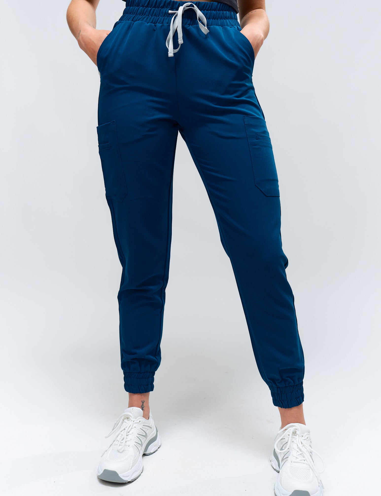 A person standing wearing blue jogger scrub pants with a drawstring waistband, paired with white sneakers.