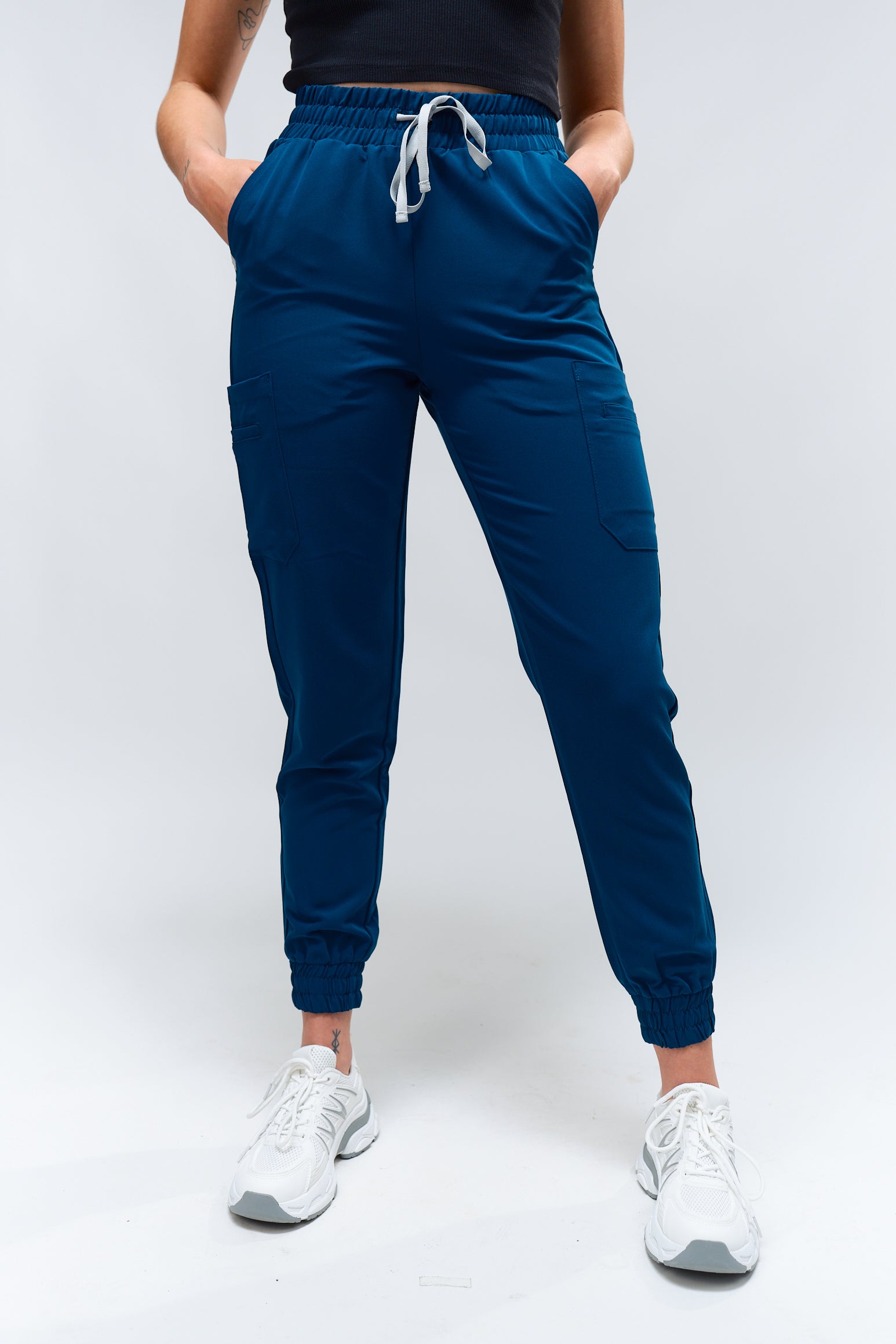 A person standing wearing blue jogger scrub pants with a drawstring waistband, paired with white sneakers.
