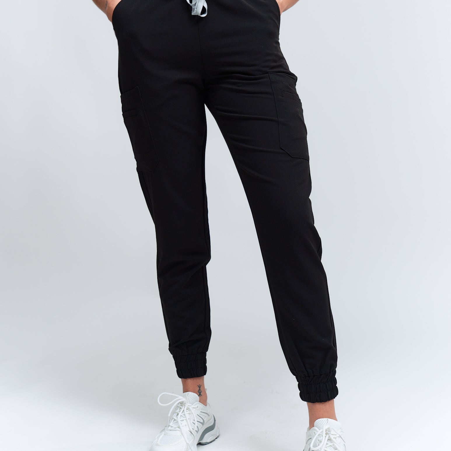 A person wearing black jogger scrub pants with multiple pockets and a drawstring waist, paired with a black scrub top and white sneakers.