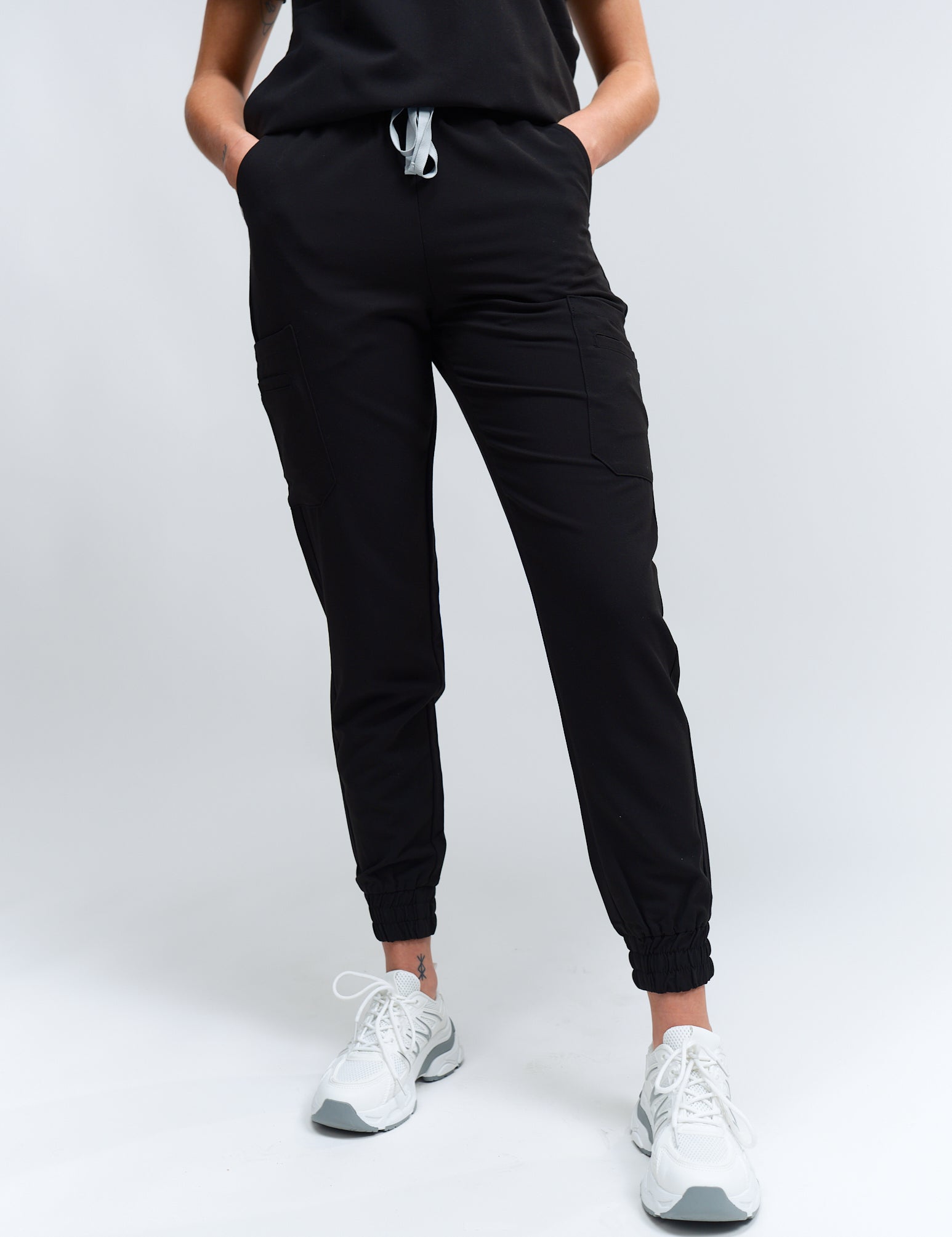 A person wearing black jogger scrub pants with multiple pockets and a drawstring waist, paired with a black scrub top and white sneakers.