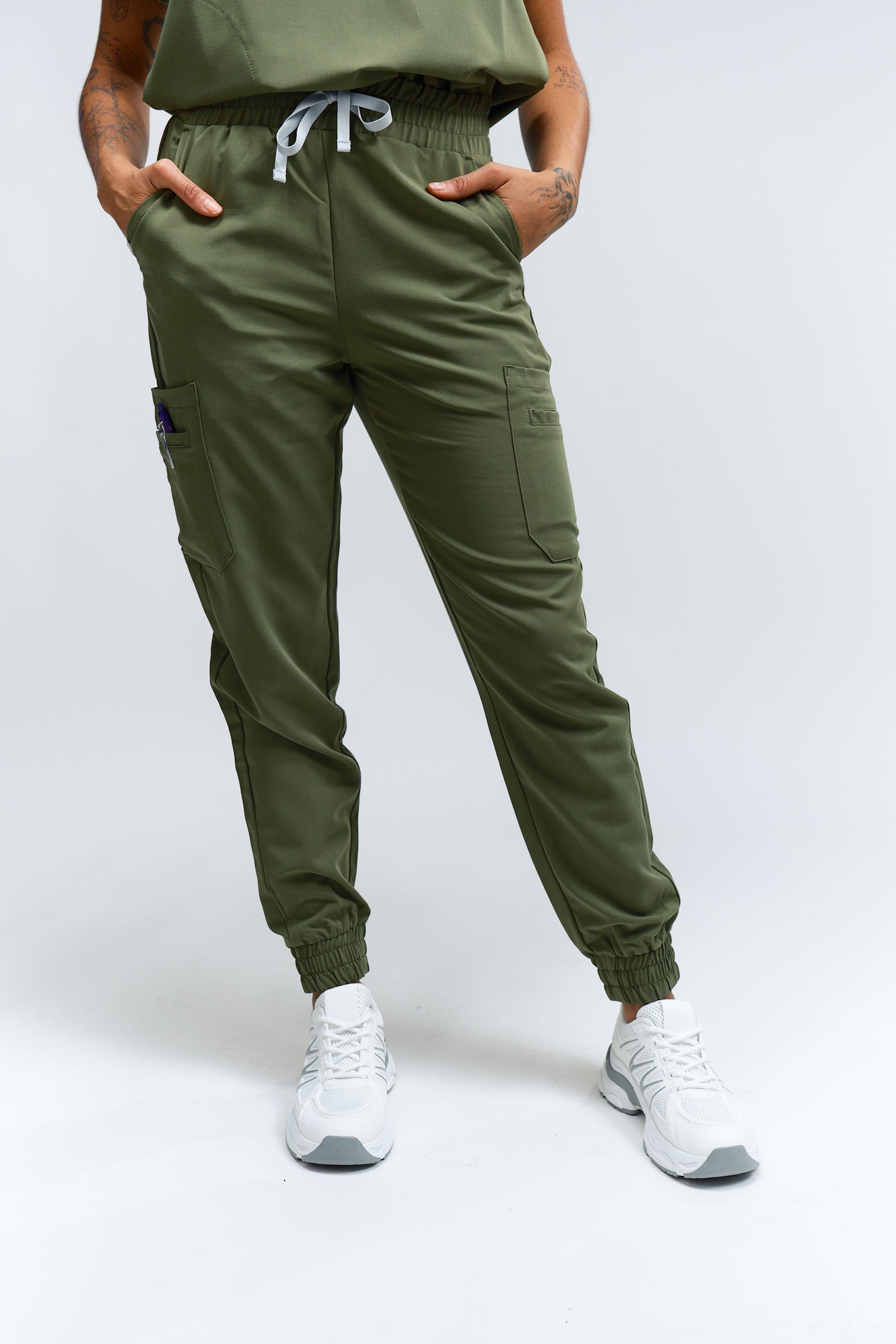 Une personne portant un pantalon de gommage vert mousse avec de multiples poches et un cordon de serrage à la taille, associé à des baskets blanches.