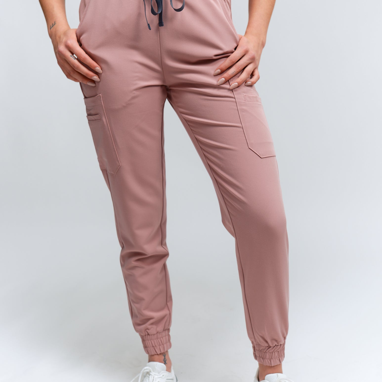 A pair of rosewood-colored jogger scrub pants with multiple pockets and a drawstring waistband, paired with white sneakers.