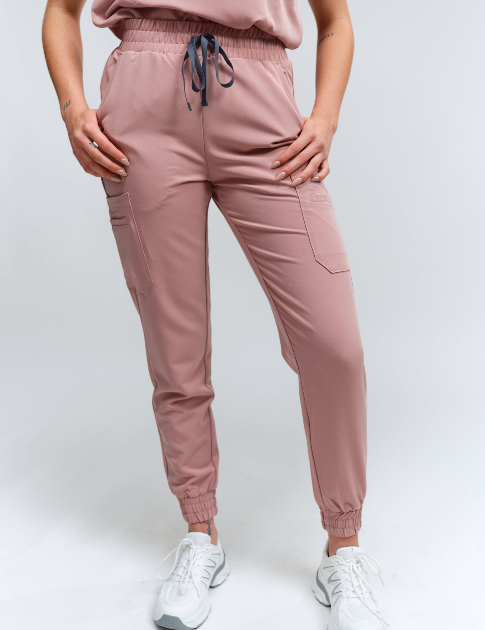 A pair of rosewood-colored jogger scrub pants with multiple pockets and a drawstring waistband, paired with white sneakers.
